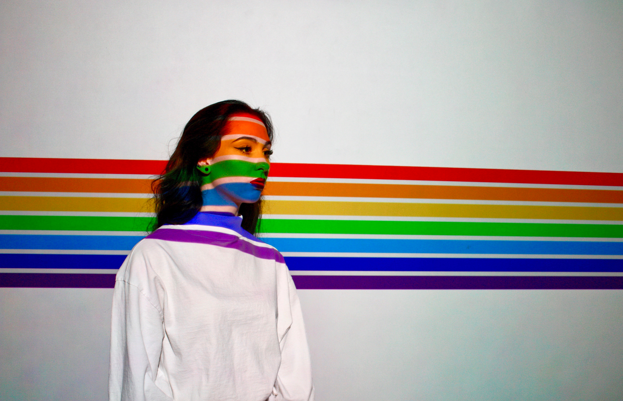 A woman in a white sweater stands against a rainbow-striped background with colorful light on her face.